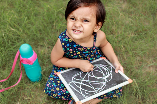 Cheerful baby girl of Indian ethnicity sitting in green grass and showing the drawing of zero.