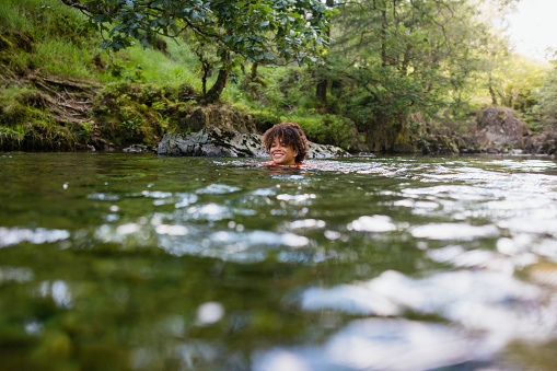 Wide shot of a woman wild swimming in the Lake District, North East of England. She is in a river, enjoying time outdoors.