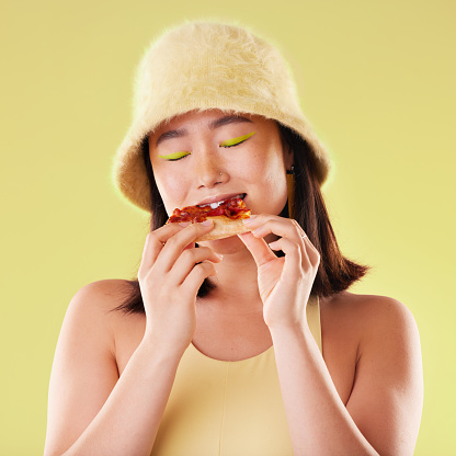 Asian girl, eating pizza and studio with fashion, happiness and retro 90s aesthetic by yellow background. Young gen z model, junk food and happy with meal, cheat snack and beauty with facial makeup