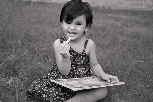 Cheerful baby girl of Indian ethnicity sitting in green grass and studying alone.