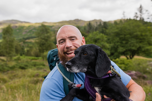 A mid adult man enjoying a day out exploring in nature in Broughton-in-Furness, Lake District. He is carrying his pet Beagador while looking at it and smiling.