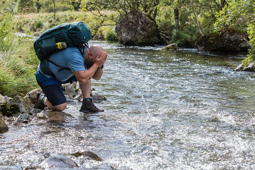 A mid adult man enjoying a day out exploring in nature in Broughton-in-Furness, Lake District. He is crouching in the river and pouring water over his face from the river to refresh himself.