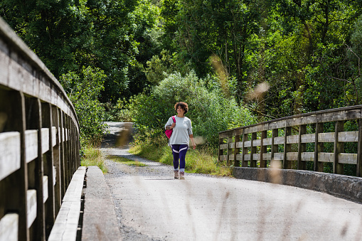 A mid adult woman enjoying a walk in nature in Broughton-in-Furness, Lake District. She is wearing a rucksack and walking over a bridge looking to the side at the view.
