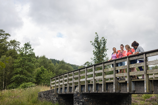 A group of female friends on a hike together in Broughton-in-Furness, Lake District. They are talking and bonding while standing on a bridge and one woman is taking a picture of the view with her mobile phone.