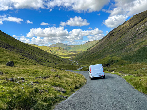 A van driving on a road through the hills in Broughton-in-Furness, Lake District. The sun is shining over the green mountain range.