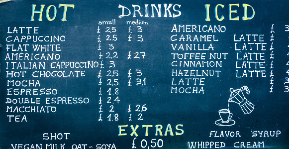 Close up color image depicting a blackboard sign at an outdoor food and drink market advertising a selection of hot drinks on offer. The selection includes different varieties of tea, hot chocolate and coffee. Focus is on the sign with the market stall defocused in the background. Room for copy space.