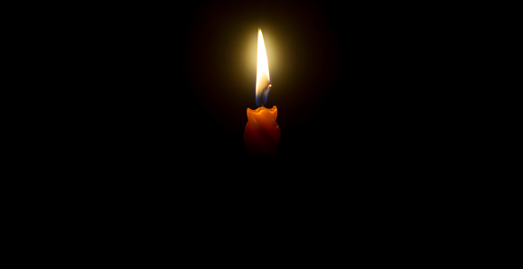 A single burning candle flame or light is glowing on an orange candle on black or dark background on table in church for Christmas, funeral or memorial service with copy space.