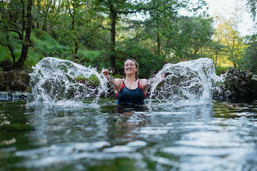 Wide shot of a woman wild swimming in the Lake District, North East of England. She is in a river, enjoying time outdoors, splashing.