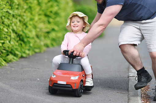 Young toddler laughing as she zooms down a residential path sitting on a toy car being pushed along by her dad.