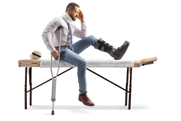 injured man with an orthopedic boot sitting on a therapy table and holding his head - physical injury men orthopedic equipment isolated on white imagens e fotografias de stock