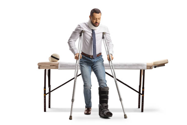 full length portrait of a man with an orthopedic boot walking with crutches in front of a therapy bed - physical injury men orthopedic equipment isolated on white imagens e fotografias de stock