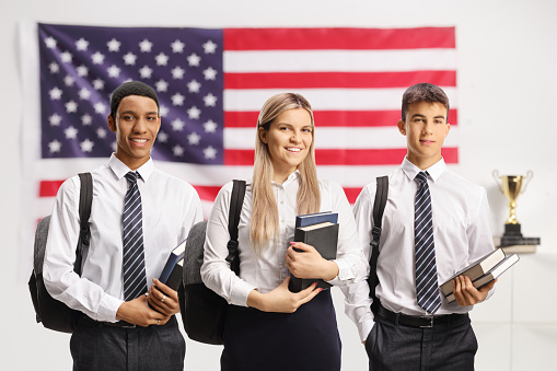 Caucasian male and female and african american male student holding books and standing in front of a USA flag