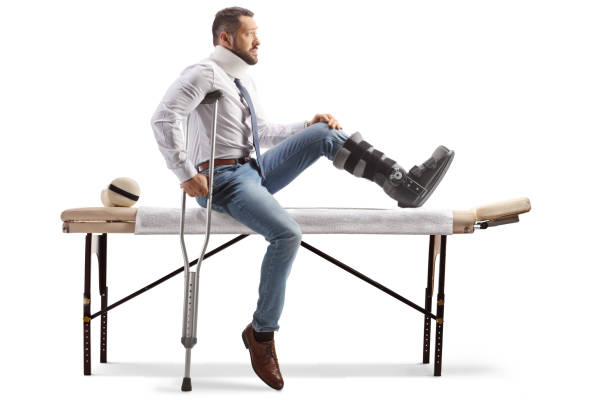 Profile shot of an injured man with an orthopedic boot and arm sling sitting on a therapy table Profile shot of an injured man with an orthopedic boot and arm sling sitting on a therapy table isolated on white background man with arm sling stock pictures, royalty-free photos & images