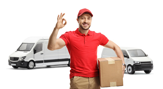 Delivery man with a package gesturing a great sign in front of transport vans isolated on white background