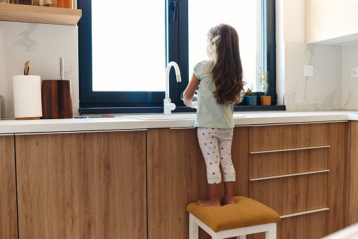Little girl drinking water from the glass in the kitchen. She is so small that she is standing on a chair.