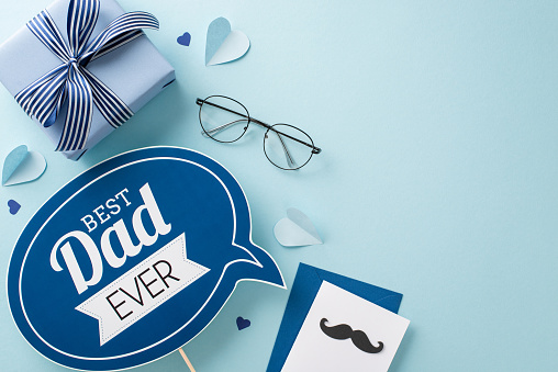 A top view flat lay for Father's Day with a greeting plaque, gift box, spectacles, paper hearts, envelope with postal on a pastel blue background with an empty space for text or advertisement