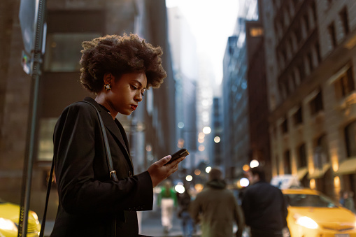 A young African-American businesswoman stands on a street corner in Wall Street, anxiously awaiting a taxi. She holds a folder filled with important documents, indicating her busy schedule, while multitasking by checking her email on her mobile phone. 
Fun fact: African-American women have been making significant strides in the business world, with a growing presence on Wall Street and a rise in entrepreneurship.