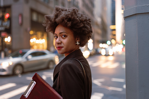 On the streets of Wall Street, a vibrant scene unfolds, featuring a young African-American woman immersed in her busy work life. With a firm grasp on her responsibilities, she eagerly scans the street, awaiting the arrival of a taxi. Holding a folder containing important documents, she skillfully manages her time by checking her email on her smartphone. 
Fun fact: Black women have been instrumental in shaping Wall Street's corporate landscape, influencing policies and advocating for equitable opportunities for all.
