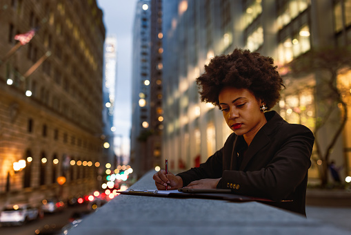 Young, stylish black businesswoman is seen multitasking as she signs documents and completes work on the go. The bustling backdrop showcases the iconic Wall Street's towering business buildings, their windows aglow with interior lights as darkness falls. 
Fun Fact: The first black woman to become a member of the New York Stock Exchange was Muriel 