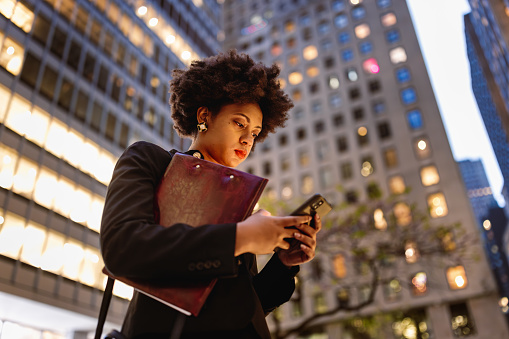 Captured in this image is a stylish, black businesswoman, embodying contemporary professionalism as she stands on the steps of a high-rise building within the heart of Wall Street. With the city's skyline aglow in the background, the illuminated windows symbolize the tireless dedication of black women who work late into the night to achieve their goals and contribute to the financial industry. 
Fun Fact: Mellody Hobson, co-CEO of Ariel Investments and a prominent black businesswoman, is known for her commitment to empowering women and promoting diversity in the workplace.