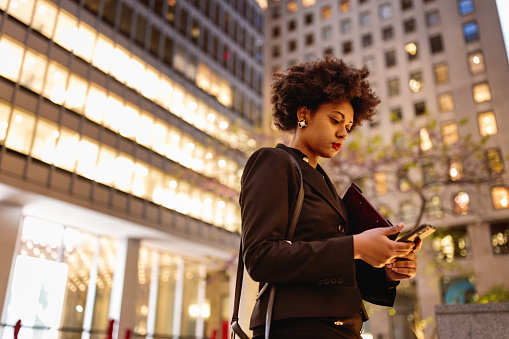 This photograph showcases a young black businesswoman at the end of her workday, positioned on the steps outside a prominent office tower. The bustling energy of Wall Street lingers in the background, emphasized by the illuminated windows of neighboring buildings. While making a phone call to secure transportation, she exemplifies the modern professional who navigates the demands of a career, even during late hours. 
Fun Fact: Black women, like Carla Harris, Vice Chairman of Wealth Management at Morgan Stanley, have made significant contributions to the finance industry by excelling in leadership roles and inspiring others.