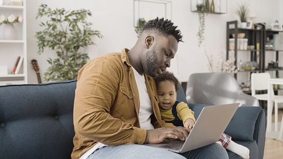 Father works on his laptop while showing his son something on the screen, paternity leave