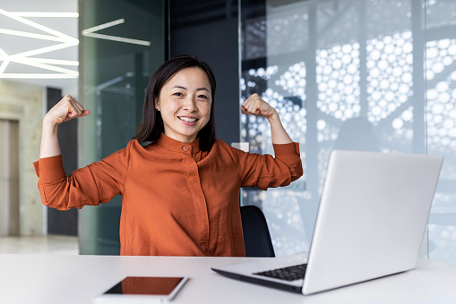 A young Asian business woman working in the office using a laptop, confidently and smilingly looking at the camera and showing a power gesture with her hands. Successfully completed work, project.