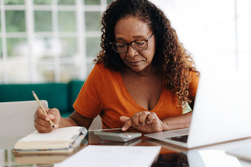 Senior woman making calculations on her retirement annuity, reviewing her pension plan and social security benefits. Elderly black woman using her financial literacy and planning skills at home.