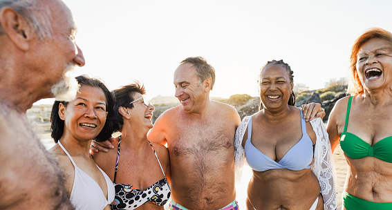Senior multiracial friend wearing swimsuit while walking on the beach - Joyful elderly lifestyle, travel, vacation and summer concept - Main focus on center people faces