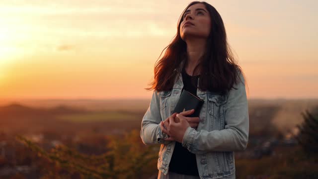 Beautiful young woman reading the bible while standing on top of a hill with a beautiful view at sunset, admiring the view and nature, relaxing alone with thoughts, slow motion