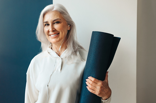 Mature woman stands holding a rolled-up yoga mat, preparing to start her yoga practice for the day. Senior woman embracing a healthy lifestyle with the addition of yoga to her fitness routine.