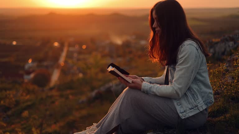 A woman reads a book in the sun. A girl is reading the Bible in the open air, holding the Bible in her hands and studying the word of God at sunset on the top of the mountain. Self-educational concep