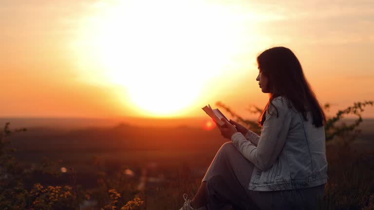 A girl reads the Bible in the open air. A woman holds a Bible in her hands and studies the word of God while sitting in a field at sunset