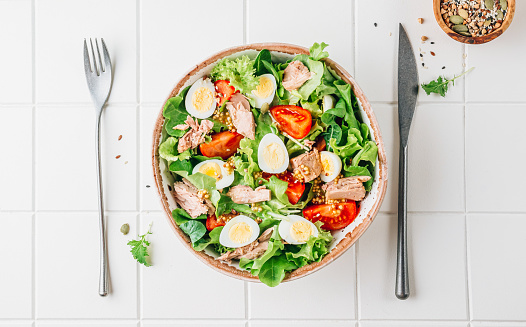 Healhty tuna salad with tomato, quail eggs, lettuce and salad dressing with olive oil, lemon juice and grain mustard. Tasty diet salad over white tiled background