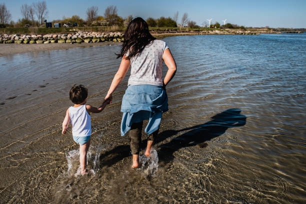 Mother and son run happily on a beach in the shallow water hand in hand. Kid running happy by the sea shore with mom conveying parent 's bound or sane parental relationship, crucial for healthy growth - Copenhagen, Denmark madre e hijo corren felices en una playa en aguas poco profundas de la mano. niño corriendo feliz por la orilla del mar con mamá transmitiendo la relación parental atada o sana de los padres, crucial para un crecimiento saludable - copenhague, dinamarca