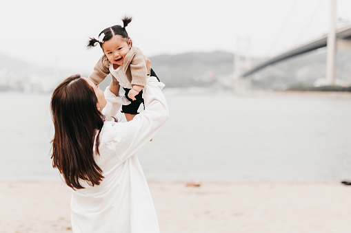 An Asian mother and her toddler are spending a beautiful day at the beach. The toddler is having a great time playing in the sand while the mother watches on with a smile.