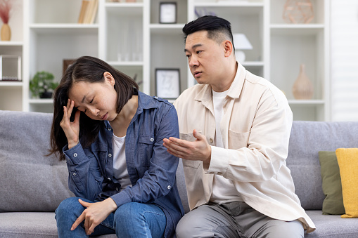 Family conflict, psychological violence, abuse. Young Asian man and woman are arguing while sitting on the couch at home. The woman listens and holds her head, the man shouts and threatens.