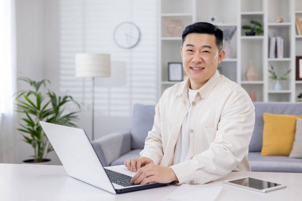 ritratto di programmatore asiatico di successo, uomo che lavora in remoto dall'ufficio di casa, uomo d'affari che sorride e guarda la fotocamera usando il laptop al lavoro seduto nel soggiorno a casa - personal organizer meeting business cyberspace foto e immagini stock