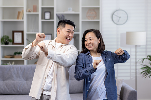 Happy Asian couple man and woman dancing together at home in daytime in living room, happy young family celebrating happy day and wedding anniversary.