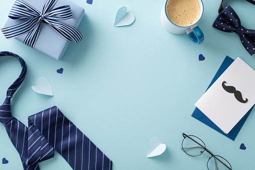 Stylish Father's Day concept. Top view shot of coffee cup, necktie, bow-tie, accessories, giftbox, and envelope with greeting card on pastel blue background with empty frame for text