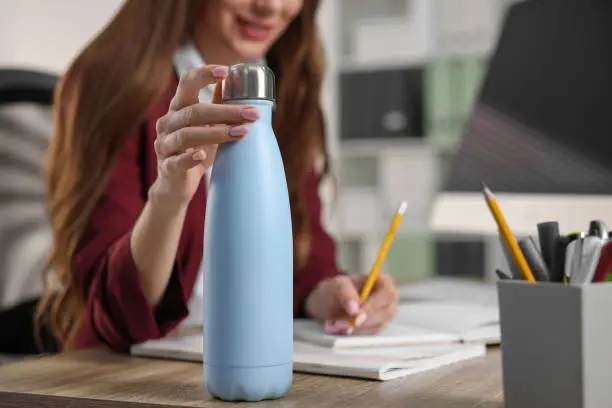 Photo of Woman holding thermos bottle at workplace, closeup