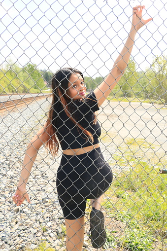 A Fijian woman using a steel mesh fence as resistance during an outdoor exercise workout.  She is wearing long, straight, brown hair, a black short t shirt, shorts and shoes.