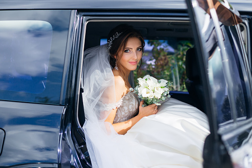 cute bride sits in a black car on the wedding day with a bouquet. Fluffy white lace dress