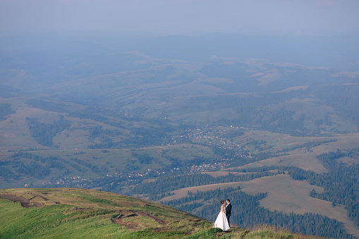 Young wedding couple standing in a field against the backdrop of mountains. The newlyweds embrace with a smile.