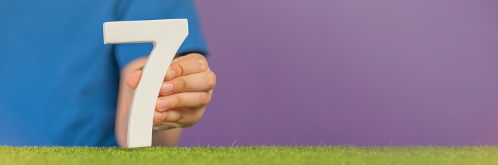 Number 7 in hand. A hand holds a white number seven on green grass and a purple background, seven percent per annum. World Health Day. April 7th. Holiday concept. High quality photo