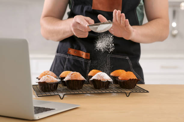 Man decorating muffins with powdered sugar while watching online cooking course via laptop in kitchen, closeup. Time for hobby