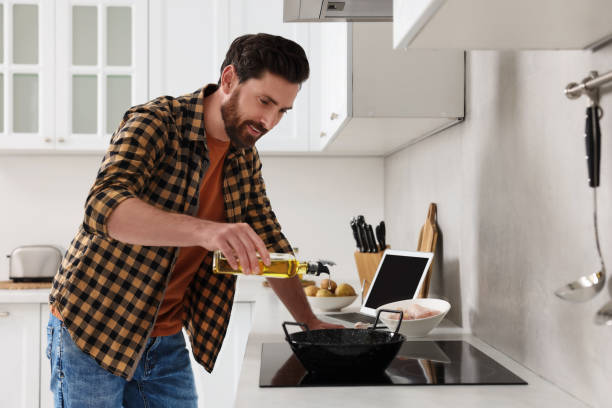Man pouring oil into frying pan while watching online cooking course via laptop in kitchen