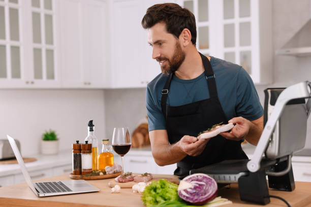 hombre preparando la cena mientras ve el curso de cocina en línea a través de una computadora portátil en la cocina - red meat fotografías e imágenes de stock