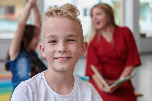 A cheerful blonde boy sits on the floor, dressed in a school uniform, smiling broadly.
