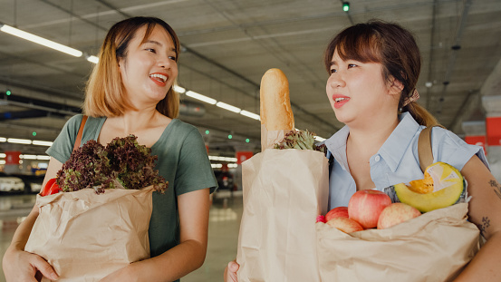 Young Asian female friends holding shopping bags full of groceries and walking in car parking. Shopping in the supermarket concept.